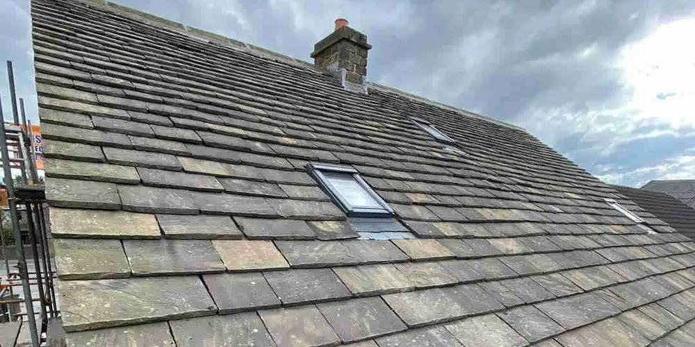 A stone slate roof with roof windows and a chimney stack, scaffold along the eaves