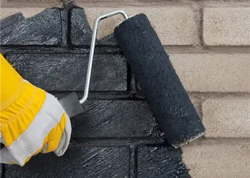 A gloved hand rolling a black damp-proof coating onto a brick wall